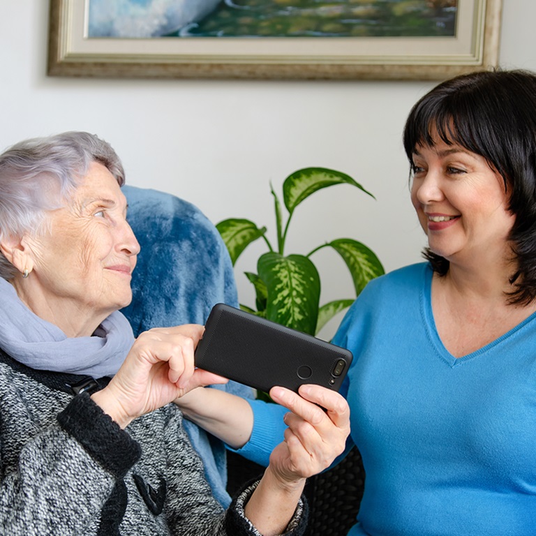 An elderly woman and a middle-aged woman sitting together indoors, with the middle-aged woman showing something on her smartphone to the elderly woman, highlighting the importance of cybersecurity in healthcare