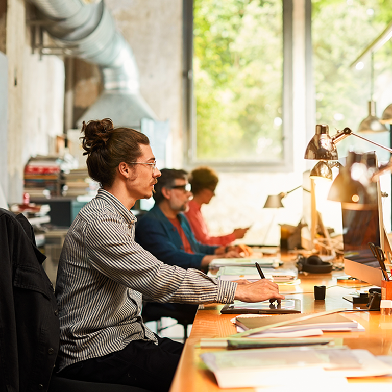 Three colleagues working at computers along a sunlit open-plan desk, used to illustrate making sense of cyber insurance.