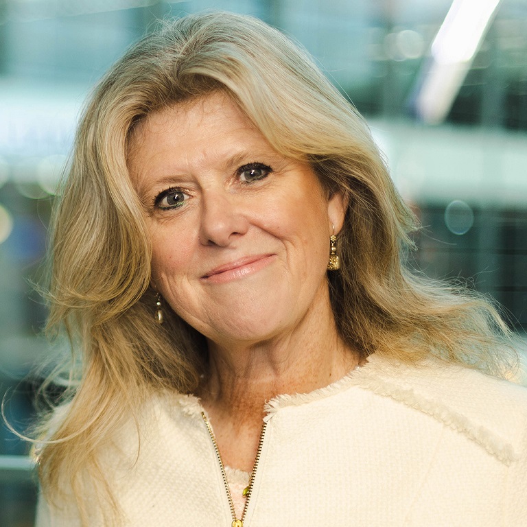 Ailsa King with long blonde hair, wearing a cream textured jacket with a zipper and gold earrings, standing in a softly blurred indoor setting.