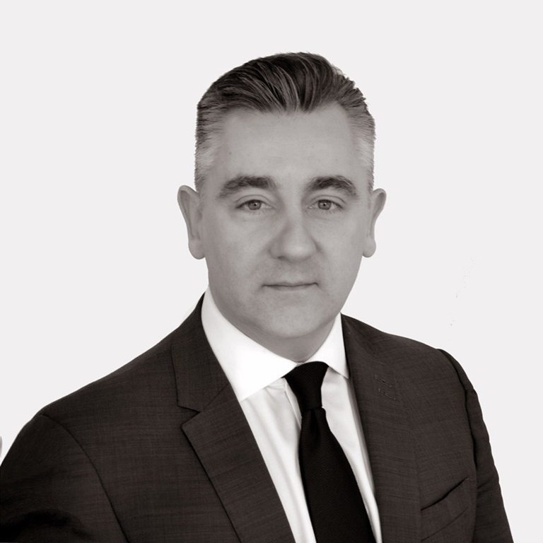 Man wearing a dark suit, white shirt, and black tie against a plain light background.