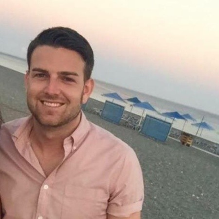 Alistair Macdonald wearing a light pink shirt standing on a beach with umbrellas and lounge chairs in the background during sunset.