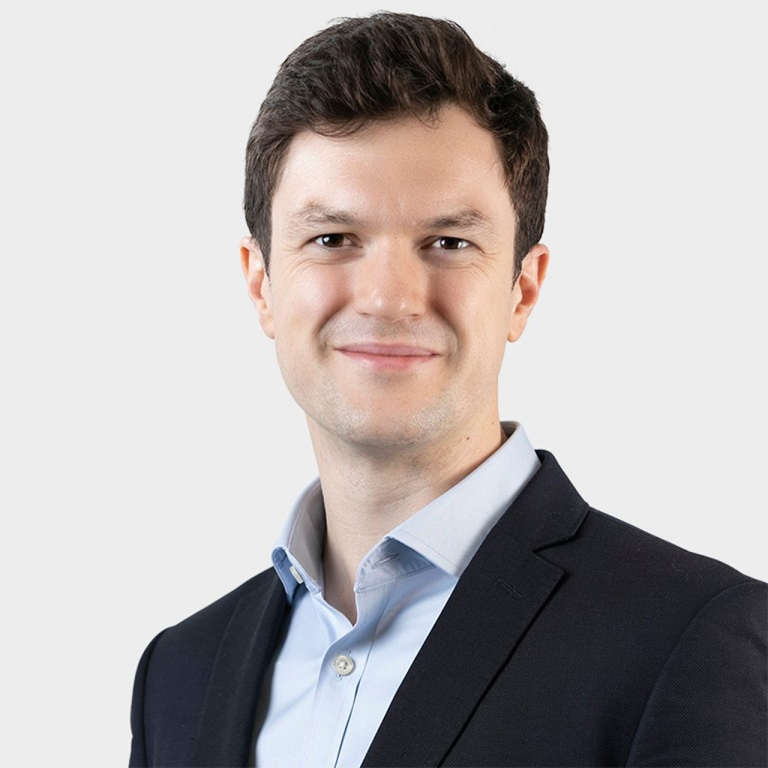 Daniel Cooper wearing a dark blazer and light blue shirt, standing against a plain background