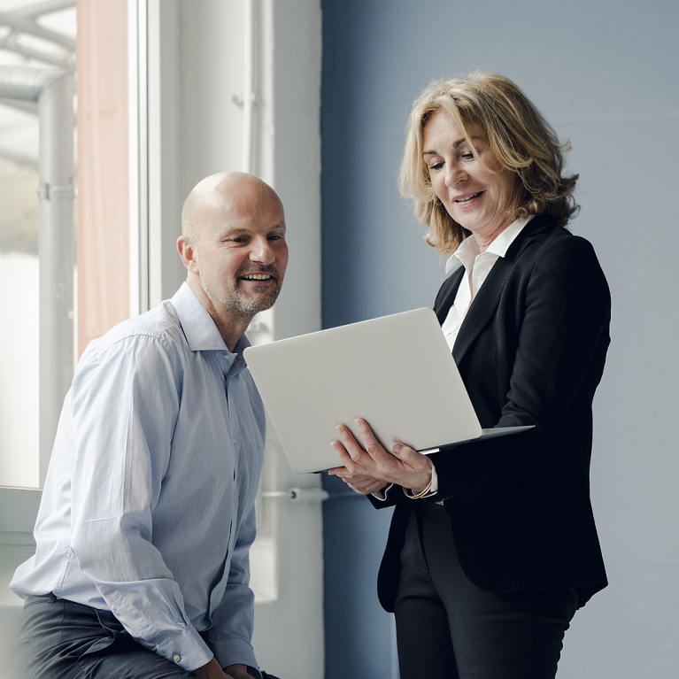 A business woman standing with a laptop showing the screen to a sitting man