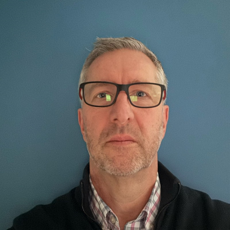 Kevin Davies wearing a black jacket over a plaid shirt, standing in front of a blue wall.
