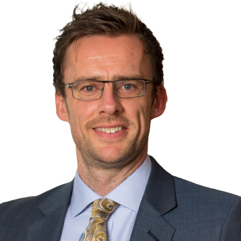 Max Houlston wearing a grey suit and patterned tie, standing confidently against a black background