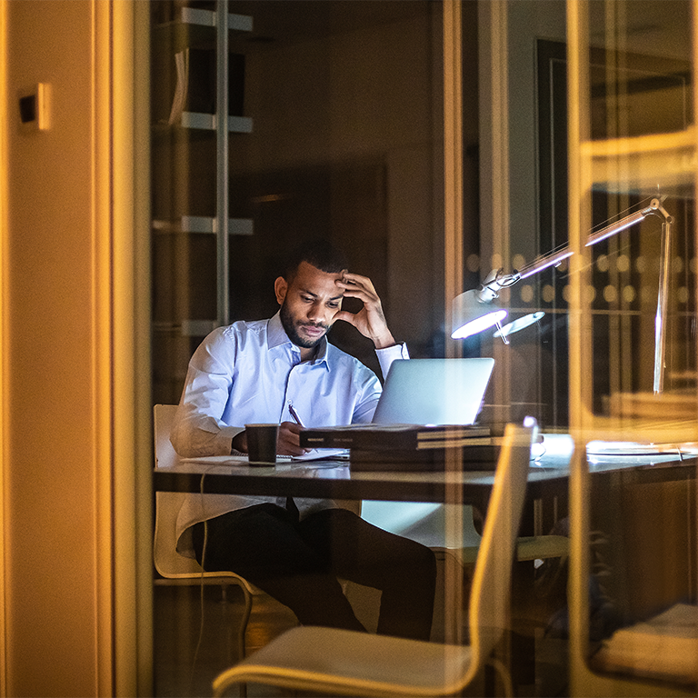 An employee working late at a desk with a laptop and documents, illuminated by a desk lamp, illustrating workplace stress claims trends