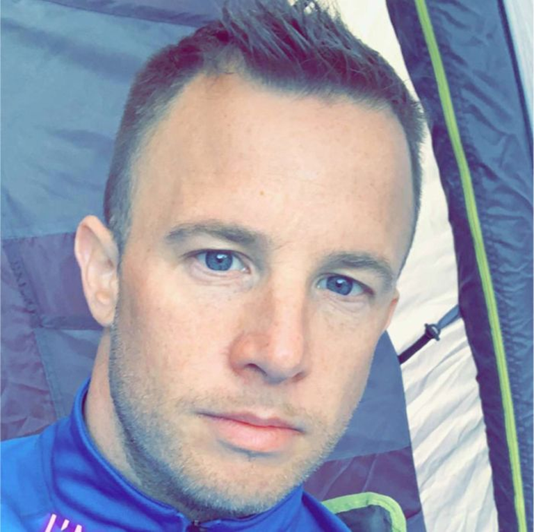 Man wearing a blue jacket sitting inside a tent with dark and light fabric panels.