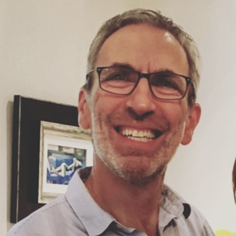 Peter Stoll smiling and wearing an open-neck shirt with a framed artwork visible in the background.