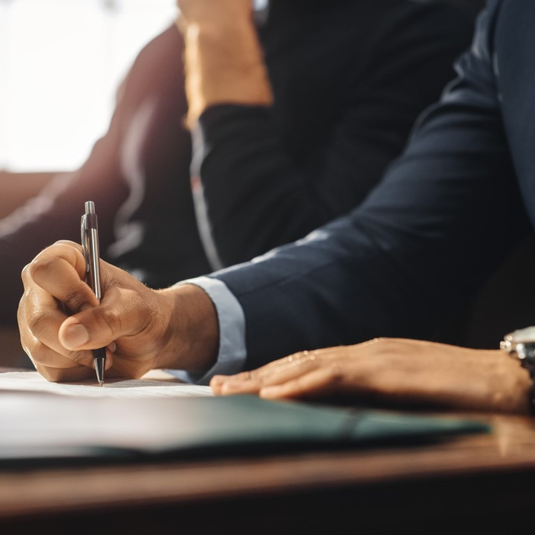 A professional person in a suit writing on documents at a desk, with a person in the background, representing solicitors' professional indemnity renewal.