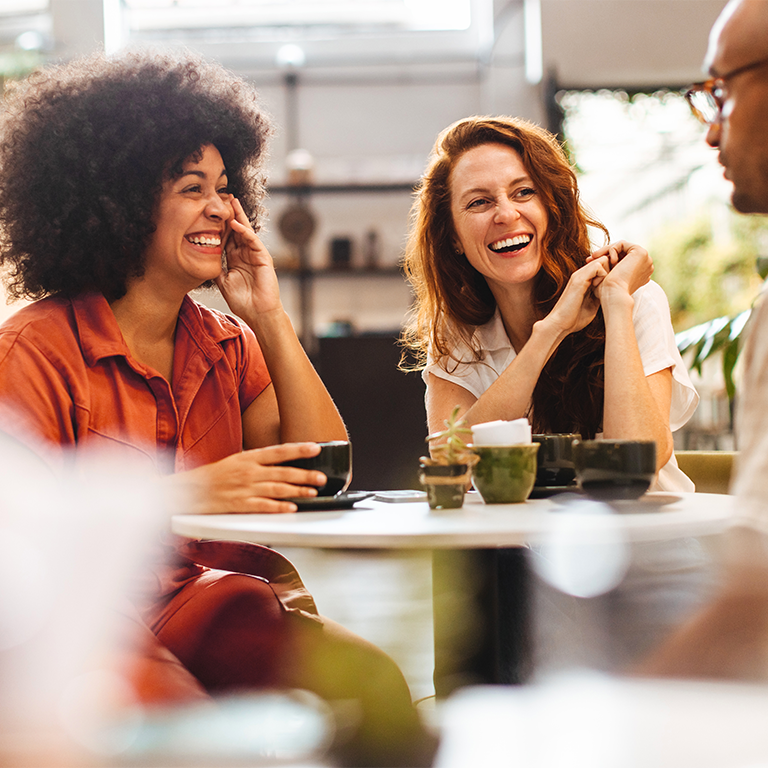 Three colleagues sit at a cafe table with cups of coffee; one person gestures while the others listen attentively in a relaxed cafe setting, illustrating psychological safety.