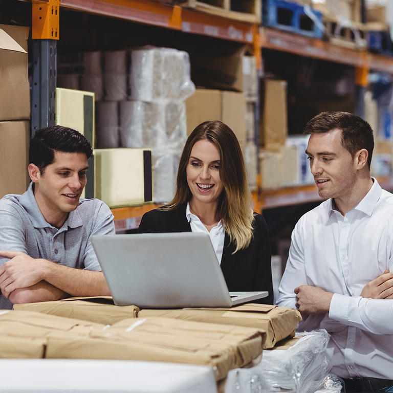 Four people gathered around a laptop in a warehouse filled with shelves of boxes and packages, discussing business operations.