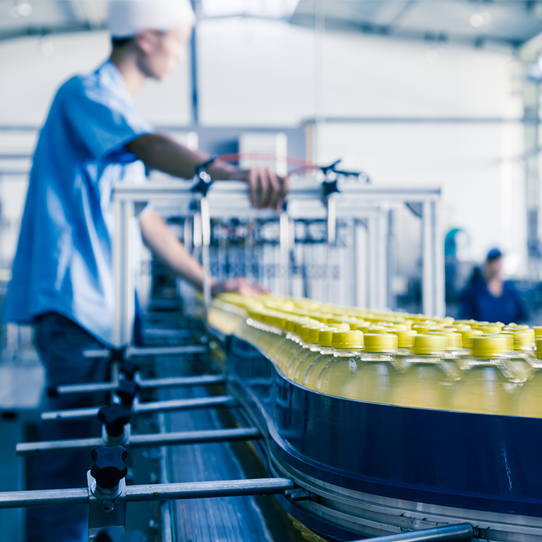 Rows of clear plastic bottles with yellow caps moving along a conveyor belt in a bright manufacturing facility with workers in the background.